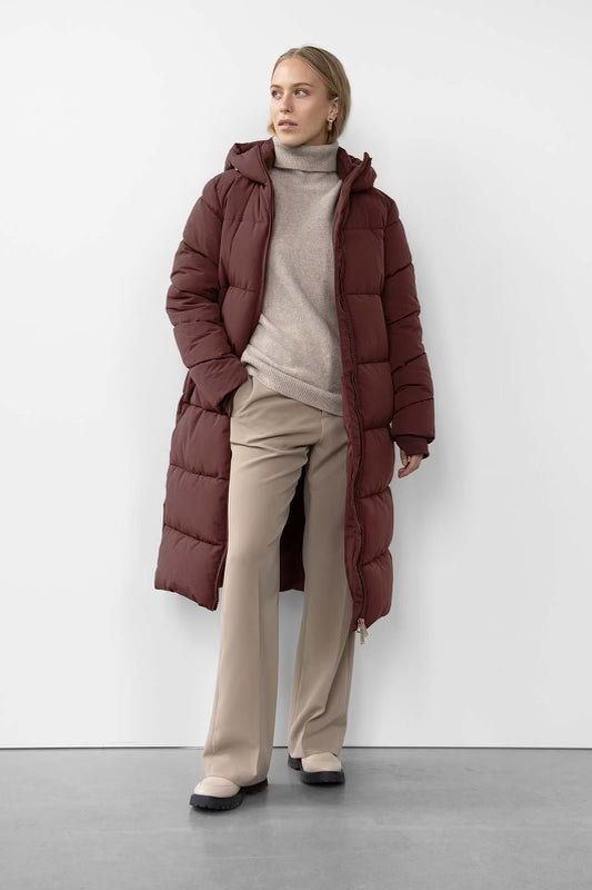 A long signature puffer coat in burgundy, insulated with fiberdown, featuring side pockets, a zipper front, and a hood for added warmth.